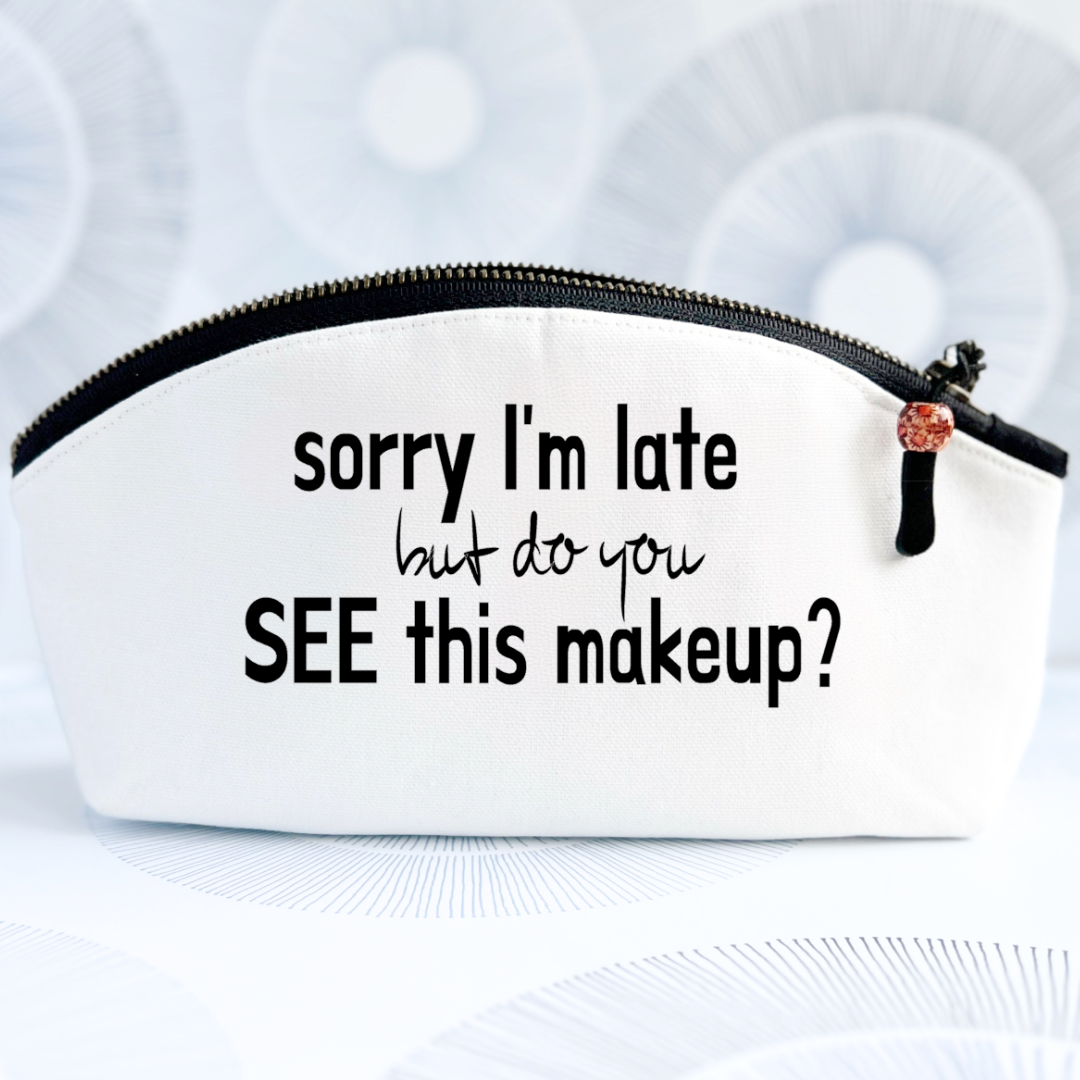 white canvas zip bag with black zipper, curved top and the words, sorry I'm late but do you see this makeup?