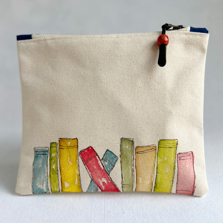 A square canvas pouch in natural cream canvas with a colorful illustration of book spines along the bottom. It has a top zipper closure with a black pull tab and a red bead accent, photographed against a neutral light gray background.