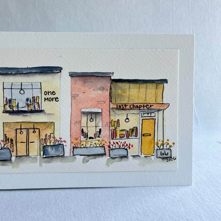 A watercolor painting of four colorful brick storefront bookstores, with the names, "Just Looking"  "One More" and "Last Chapter."  Displayed on top of a flower-painted book.