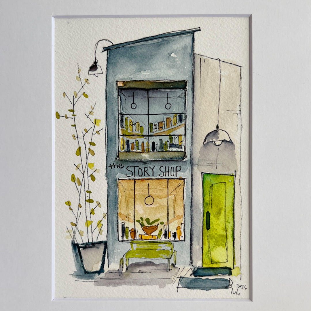 An original watercolor painting depicting a cozy, tall, narrow bookstore with the name "the Story Shop," with shelves, a green door, a bench, and a potted plant, presented in a white frame against a light gray background.