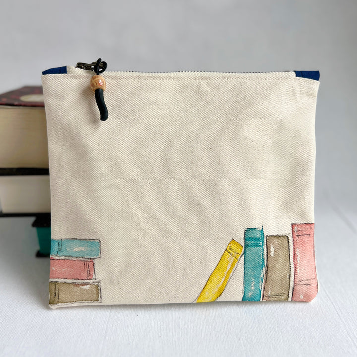 A square canvas pouch in natural cream canvas with a watercolor-style illustration of colorful book spines along the bottom. The pouch has a top zipper with a black cord pull and a wooden bead accent.