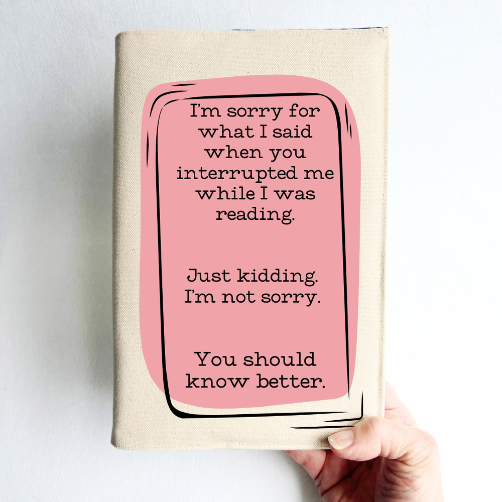 a hand holding a book covered with a linen-color reusable, readjustable book cover.  A pink square background has the words, 'I'm sorry for what I said when you interrupted me while I was reading.  Just kidding. I'm not sorry. You should know better.' in black lettering.