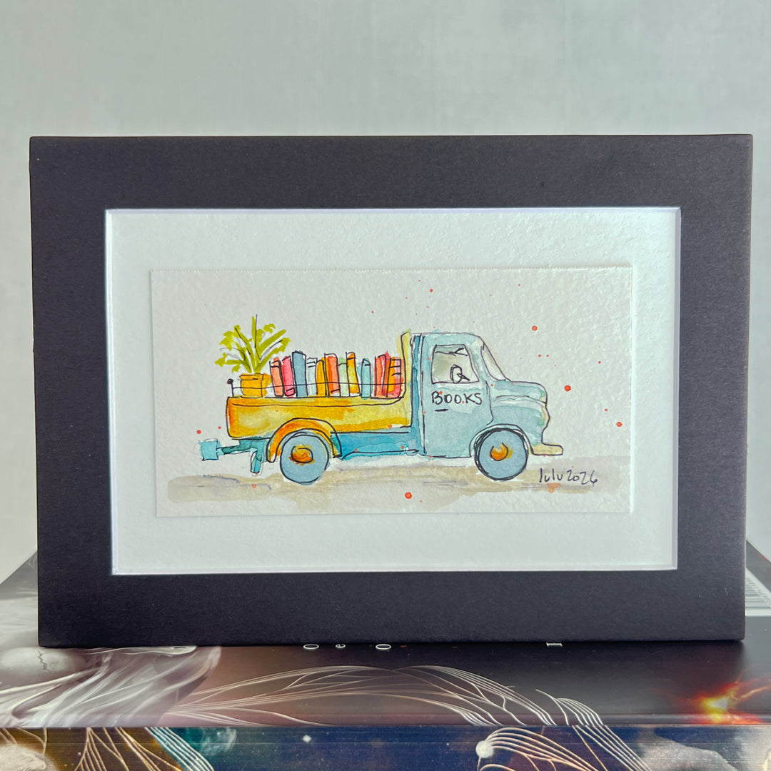 A matted watercolor art print featuring a light blue vintage truck carrying colorful books and a green plant, displayed in a black frame with a white mat on a tabletop.