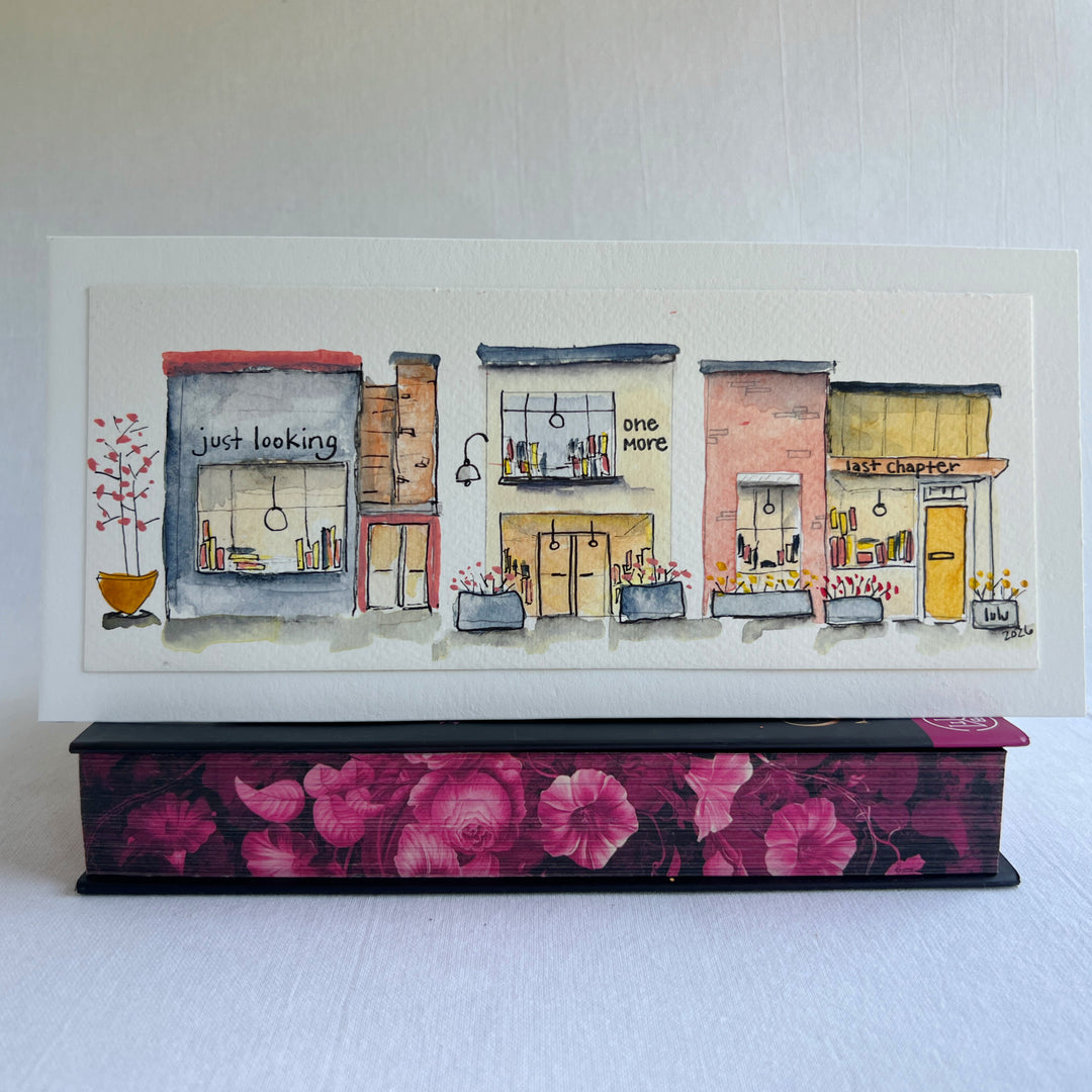 A watercolor painting of four colorful brick storefront bookstores, with the names, "Just Looking"  "One More" and "Last Chapter."  Displayed on top of a flower-painted book.