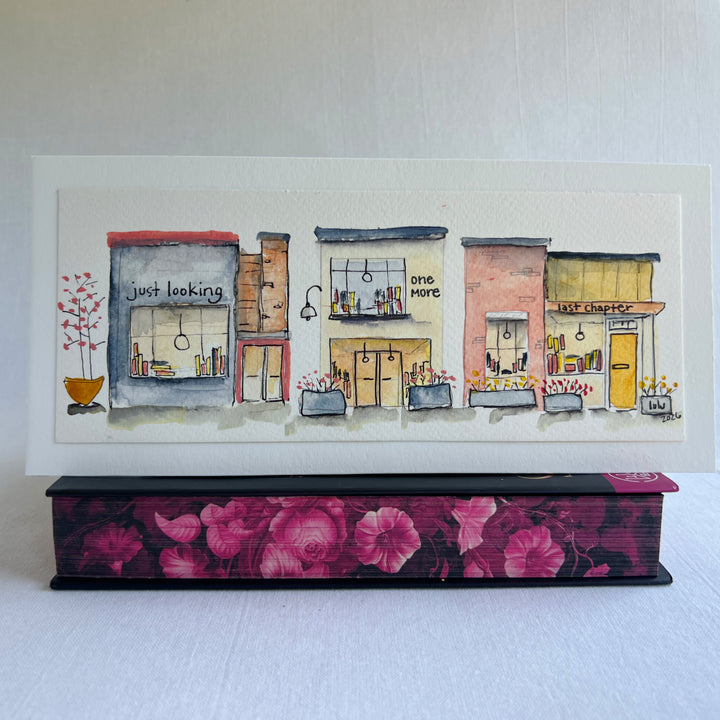 A watercolor painting of four colorful brick storefront bookstores, with the names, "Just Looking"  "One More" and "Last Chapter."  Displayed on top of a flower-painted book.