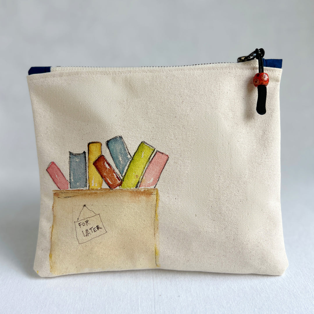 A canvas zippered pouch with a watercolor illustration of colorful books in a box labeled 'FOR LATER'. Several books are tucked inside the pouch, which is photographed against a light gray background.