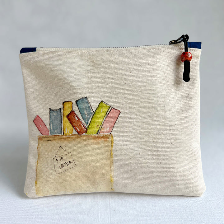 A canvas zippered pouch with a watercolor illustration of colorful books in a box labeled 'FOR LATER'. Several books are tucked inside the pouch, which is photographed against a light gray background.