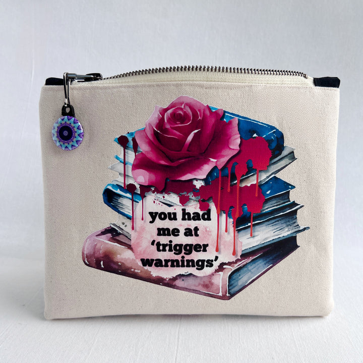 square zippered bag made of canvas, with a design depicting a stack of books, a rose and blood drips, with the words, "you had me at trigger warnings."