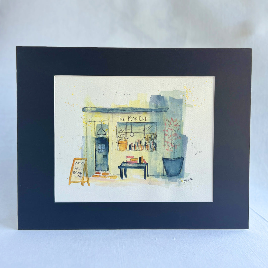 A watercolor painting of a mystery bookstore storefront labeled 'The Book End,' with a dark door, window display of books, a small table with books, potted plants, and a chalkboard sign reading 'Books Save Everything.' The artwork is painted on a white paper background with a dark navy border around it.