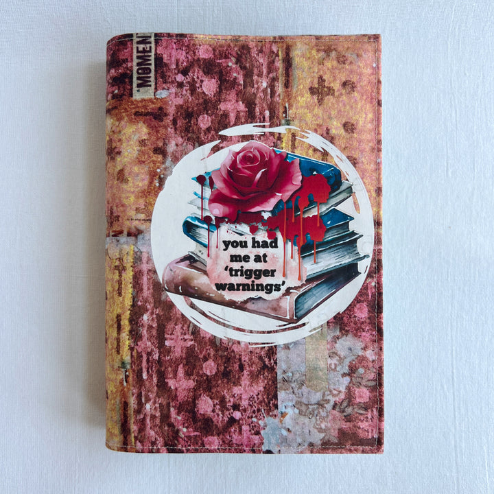 a book covered in a colorful fabric reusable book cover in colors of pinks and orange. A white circle on the front contains an original design with a stack of books, a rose, blood drips and the words, 'you had me at trigger warnings' finishes the look.  Meant for readers of dark romance.