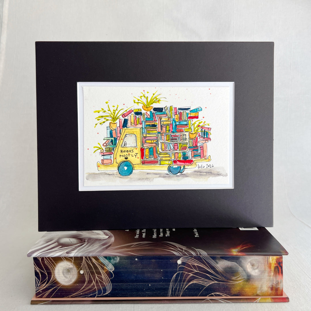A framed art print featuring a whimsical watercolor illustration of a yellow book truck loaded with books, displayed on top of a decorative book. The print is matted in a black frame against a light gray background.