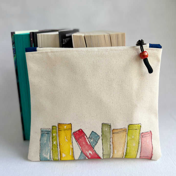 A canvas zippered pouch with colorful painted book spines in watercolor design on the front, shown with books in the background against a light gray backdrop.