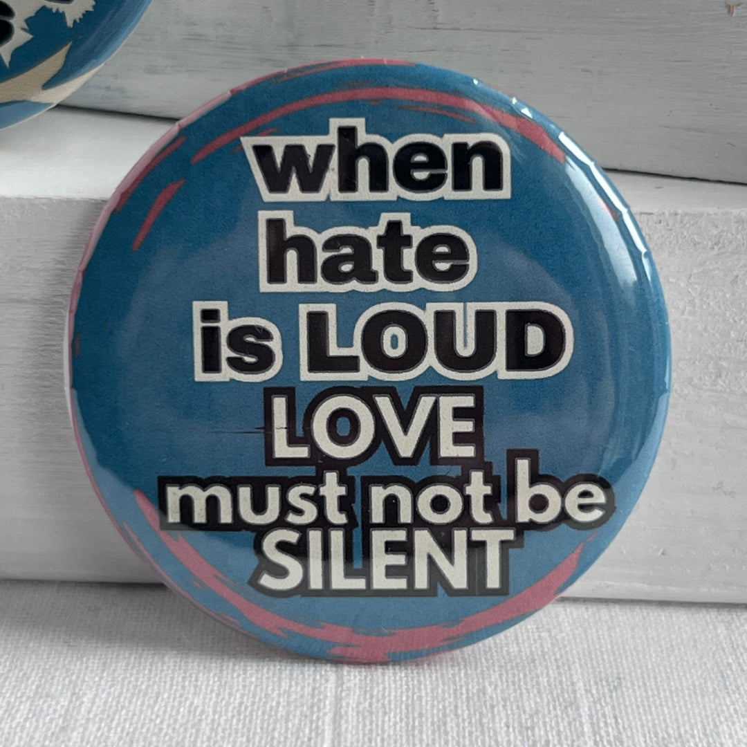 a round pinback button with a blue background and streaks of read, with the words, "when hate is LOUD, LOVE must not be SILENT."