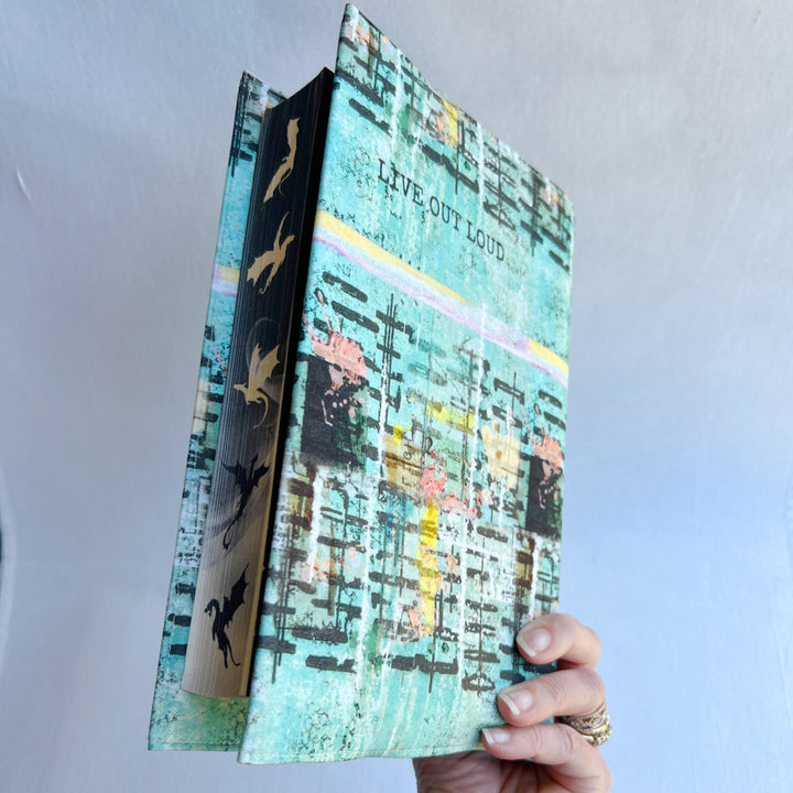 a hand holding a book covered in a vibrant blue green fabric reusable book cover with the words, 'live out loud' printed on the top.