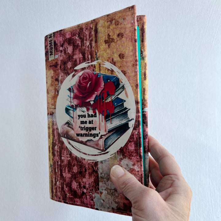 a hand holding a book covered in a colorful fabric reusable book cover in colors of pinks and orange. A white circle on the front contains an original design with a stack of books, a rose, blood drips and the words, 'you had me at trigger warnings' finishes the look.  Meant for readers of dark romance.