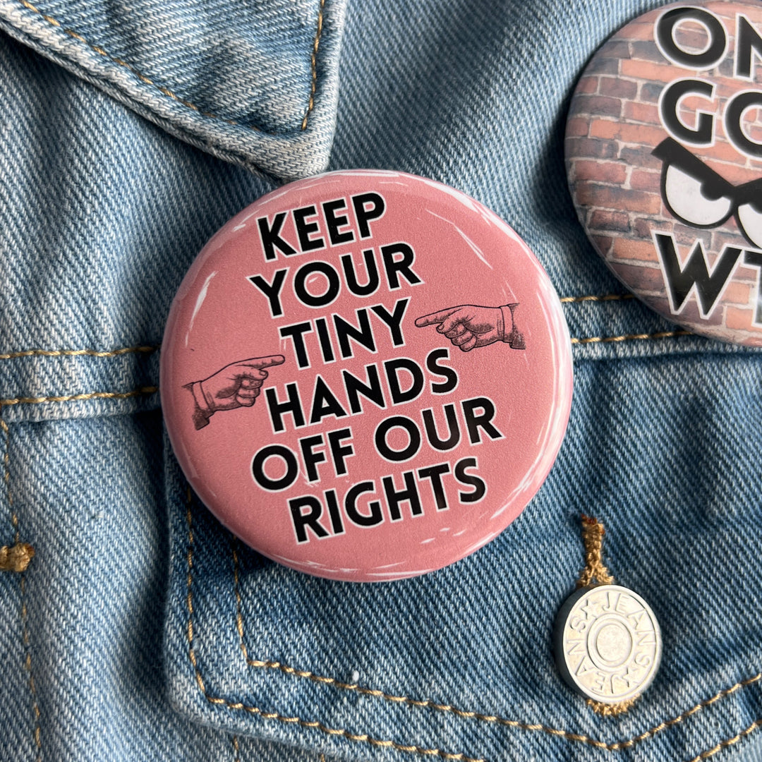 round pinback button with a pink background and the words, "Keep your tiny hands off our rights." A pair of hands in black point at the words. Pinned on a jean jacket for display.