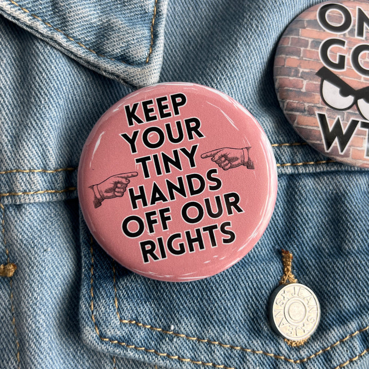 round pinback button with a pink background and the words, "Keep your tiny hands off our rights." A pair of hands in black point at the words. Pinned on a jean jacket for display.