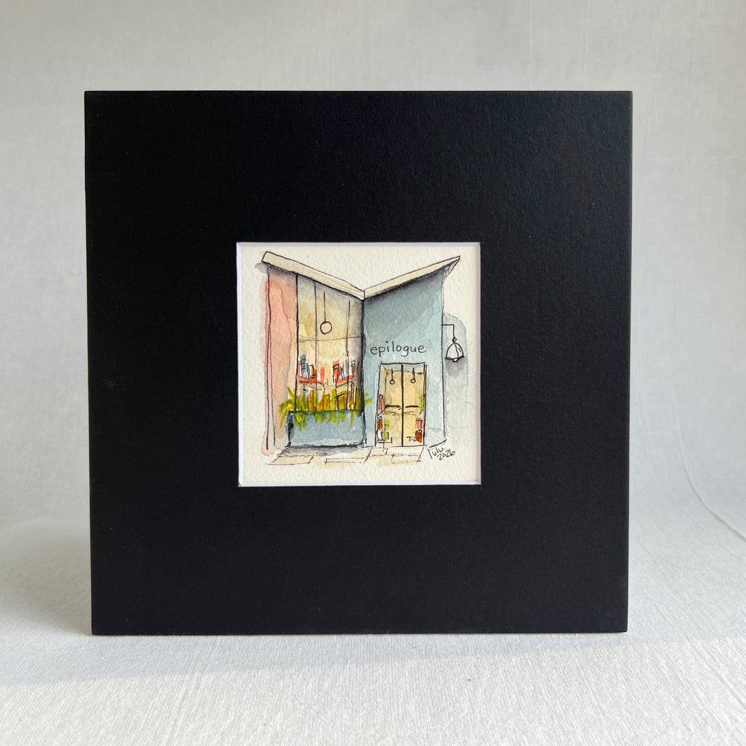 A matted watercolor painting of a cozy midcentury modern bookshop storefront with the store name  'EPILOGUE' above a window filled with books, a glass door, and a hanging lamp, shown on a white mat and white frame.