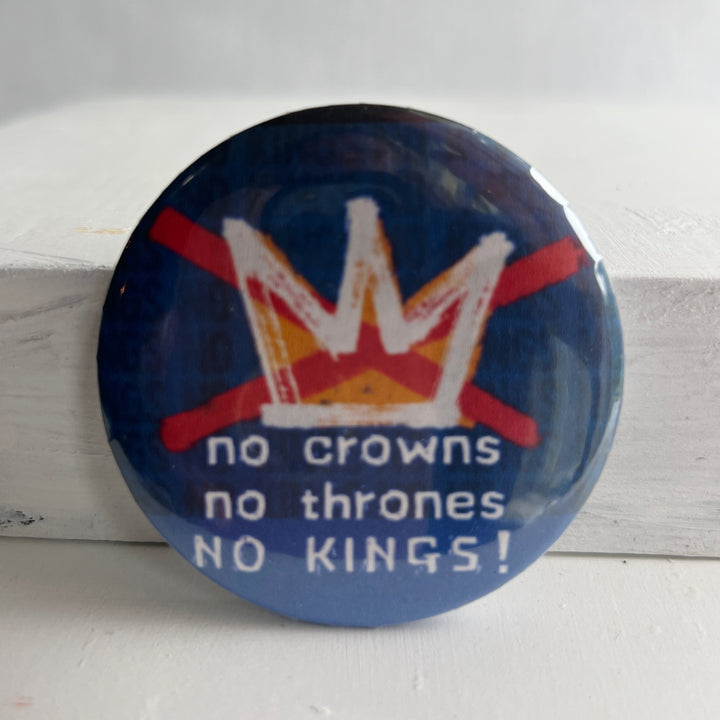 a round, blue pinback button with a white and gold crown and red slashes over it and the words, 'no crowns no thrones NO KINGS!'