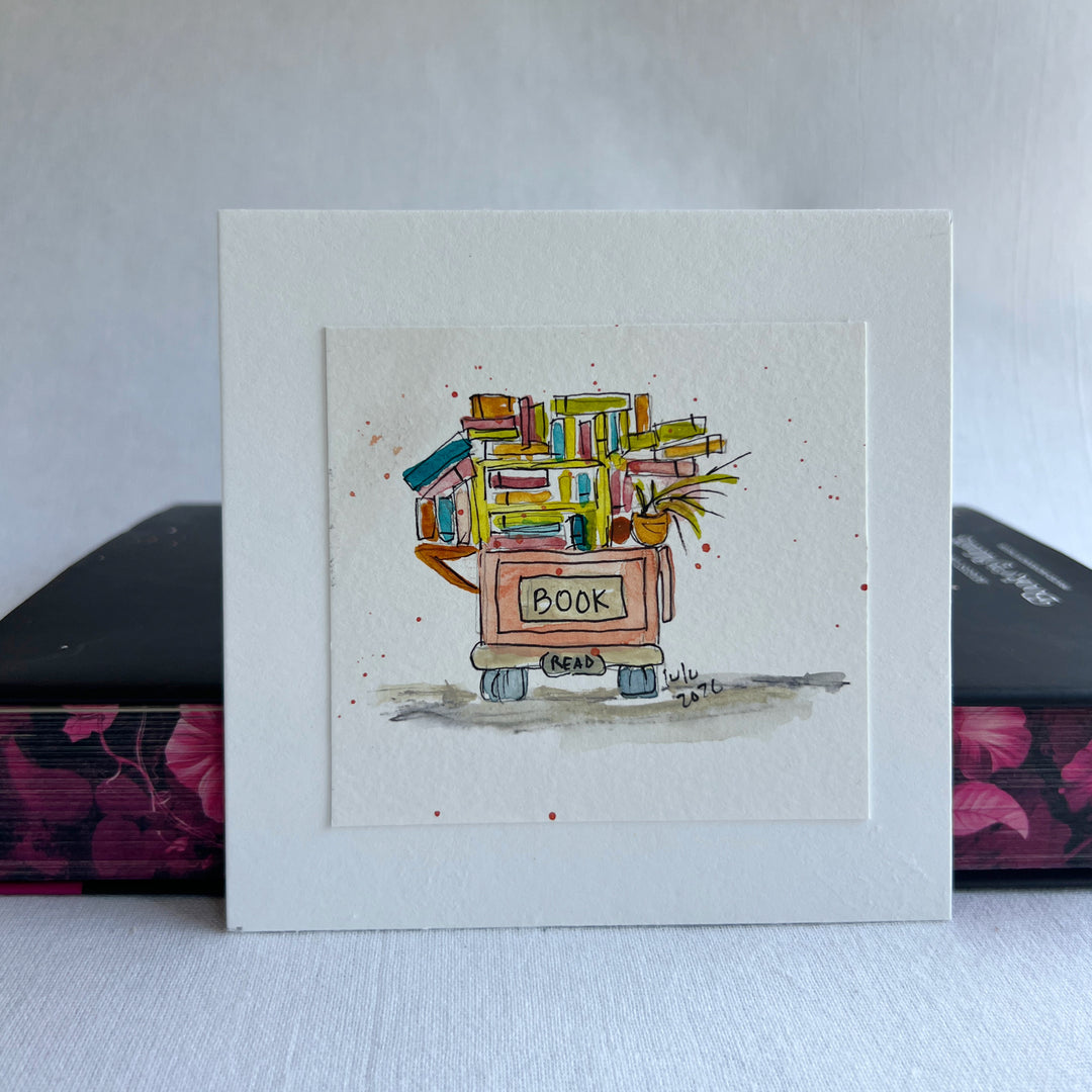 A square watercolor art print depicting a small book cart overfilled with colorful books and a potted plant, with the word “BOOK” written on the front.