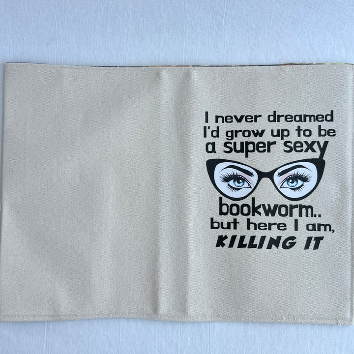 a reusable linen-color canvas book cover, with the words, 'I never dreamed I'd grow up to be a super sexy bookworm, but here I am killing it.' A pair of eyes with long lashes behind cat eye glasses completes the design.