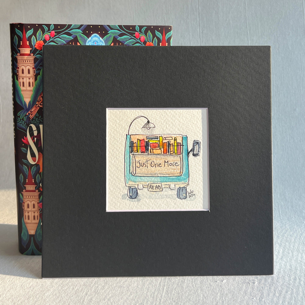A square original watercolor art print depicting the back end of a blue pickup truck loaded with colorful books and a wonky reading light, with the words, "Just One More' and a license plate with the word 'Read'  Artwork is propped in front of a colorful book.