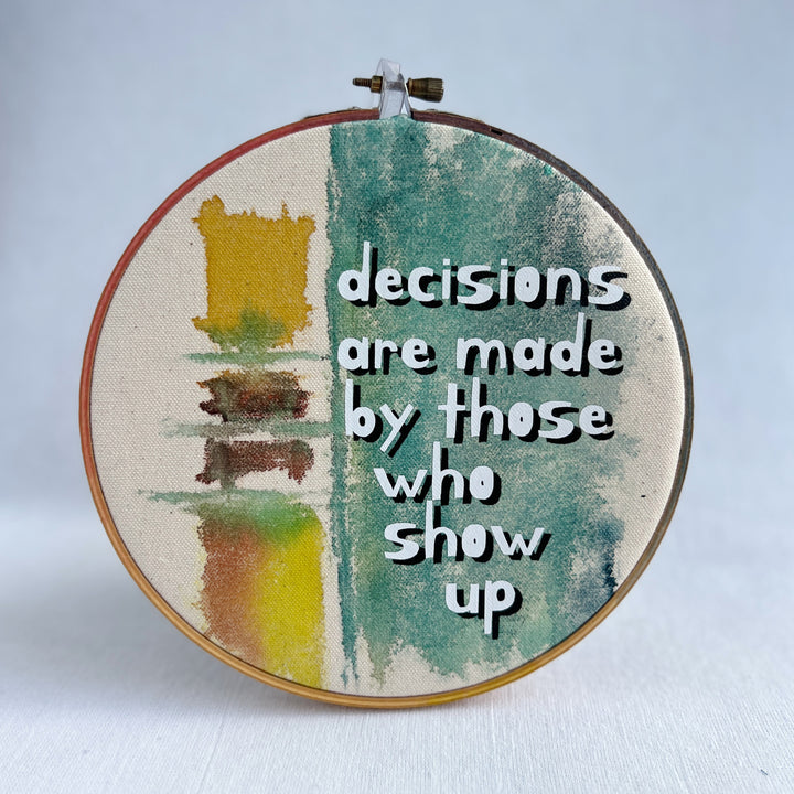 seven inch painted hoop with painted fabric in colors of yellow and mayan blues, with the words, decisions are made by those who show up.