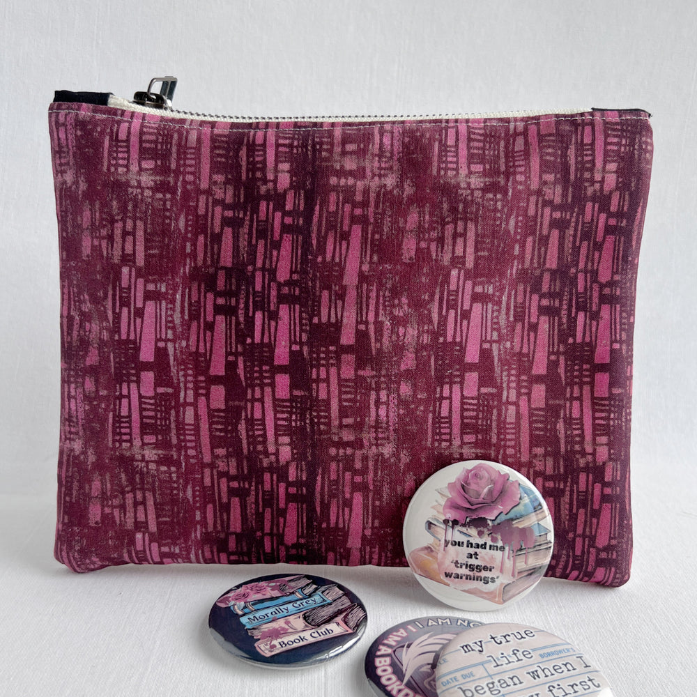 square zip bag in colors of pink and deep maroon squares, with some colorful pinback buttons scattered in front.