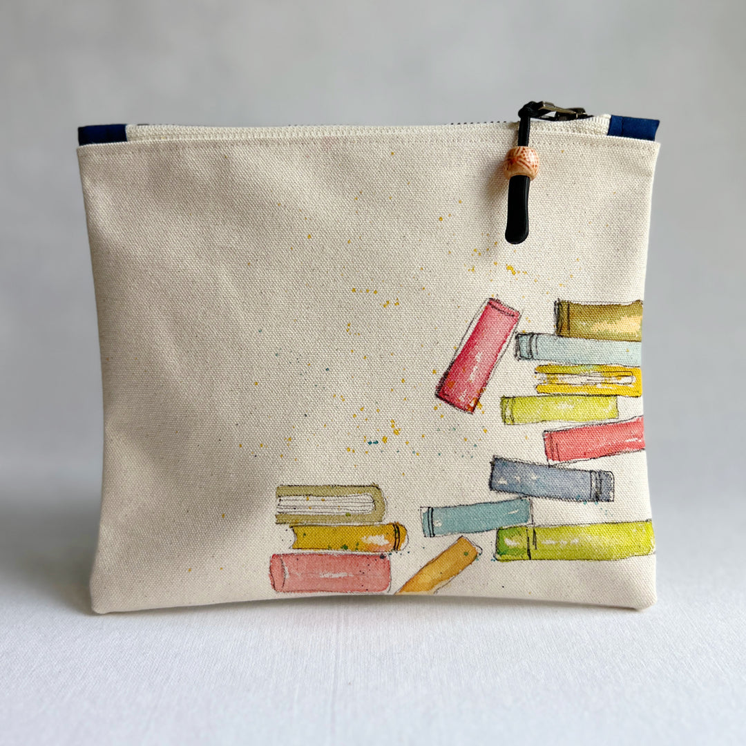 An off-white canvas zippered pouch featuring a watercolor illustration of colorful book stacks on the front, with a black zipper and pull tab.