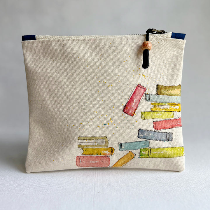 An off-white canvas zippered pouch featuring a watercolor illustration of colorful book stacks on the front, with a black zipper and pull tab.