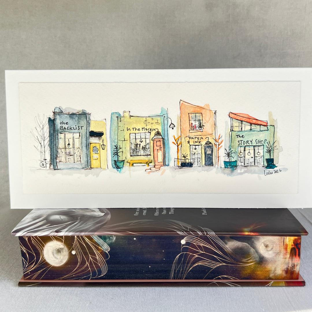 A horizontal watercolor art print depicting four whimsical bookstores with names including 'The Backlist', 'In the Margin, 'Paper & Ink', and 'The Story Shop'. The print is displayed on a textured light gray surface against a neutral gray background.