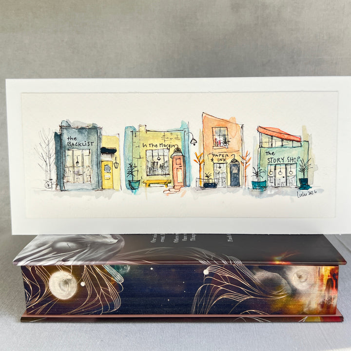 A horizontal watercolor art print depicting four whimsical bookstores with names including 'The Backlist', 'In the Margin, 'Paper & Ink', and 'The Story Shop'. The print is displayed on a textured light gray surface against a neutral gray background.