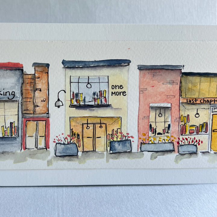 A watercolor painting of four colorful brick storefront bookstores, with the names, "Just Looking"  "One More" and "Last Chapter."  Displayed on top of a flower-painted book.