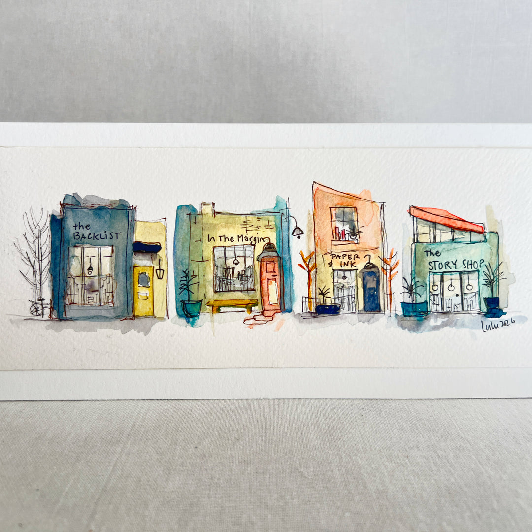 A horizontal watercolor art print depicting four whimsical bookstores with names including 'The Backlist', 'In the Margin, 'Paper & Ink', and 'The Story Shop'. The print is displayed on a textured light gray surface against a neutral gray background.