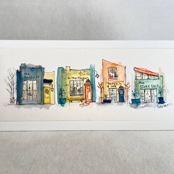 A horizontal watercolor art print depicting four whimsical bookstores with names including 'The Backlist', 'In the Margin, 'Paper & Ink', and 'The Story Shop'. The print is displayed on a textured light gray surface against a neutral gray background.