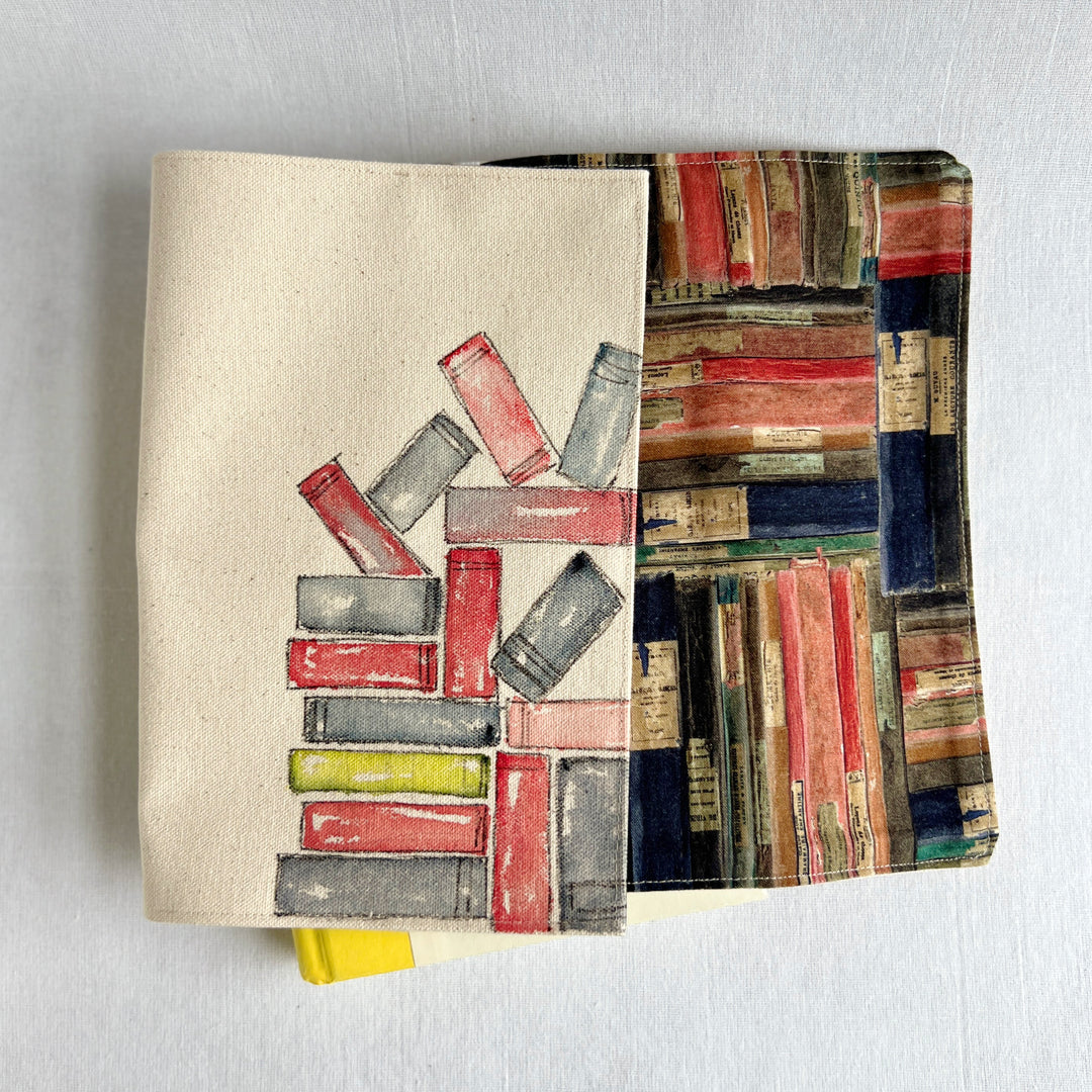 A rectangular canvas book cover featuring a watercolor illustration of stacked books in red, gray, and yellow on a beige background for the outside, and colorful book fabric for the inside.