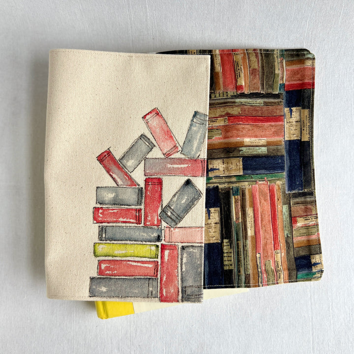 A rectangular canvas book cover featuring a watercolor illustration of stacked books in red, gray, and yellow on a beige background for the outside, and colorful book fabric for the inside.