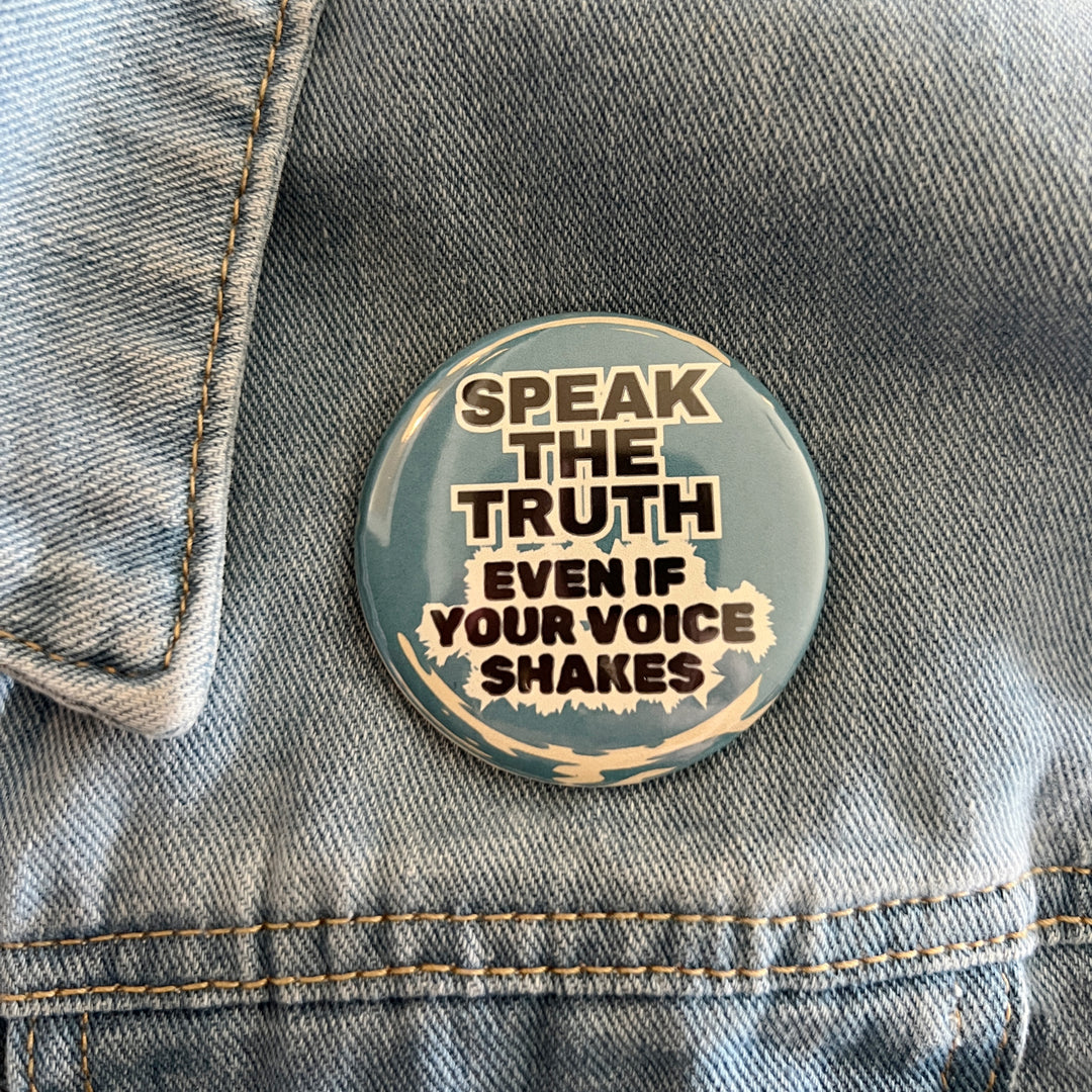 round pinback button with a blue background and the words, 'speak the truth even if your voice shakes' in black lettering bordered in white and pinned to a jean jacket for display