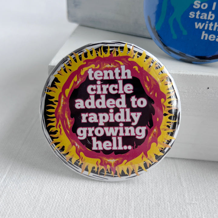 round pinback button with a black background, yellow and red flames surrounding the words, "tenth circle added to rapidly growing hell."