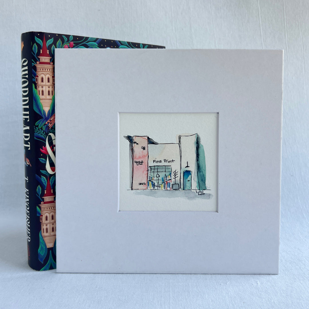 A square white art print or matted sketchbook cover featuring a small hand-drawn illustration of a building labeled 'Fine Art West.' A colorful book with a tower illustration is partially visible behind it on a light gray background.