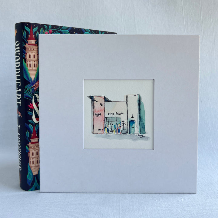 A square white art print or matted sketchbook cover featuring a small hand-drawn illustration of a building labeled 'Fine Art West.' A colorful book with a tower illustration is partially visible behind it on a light gray background.