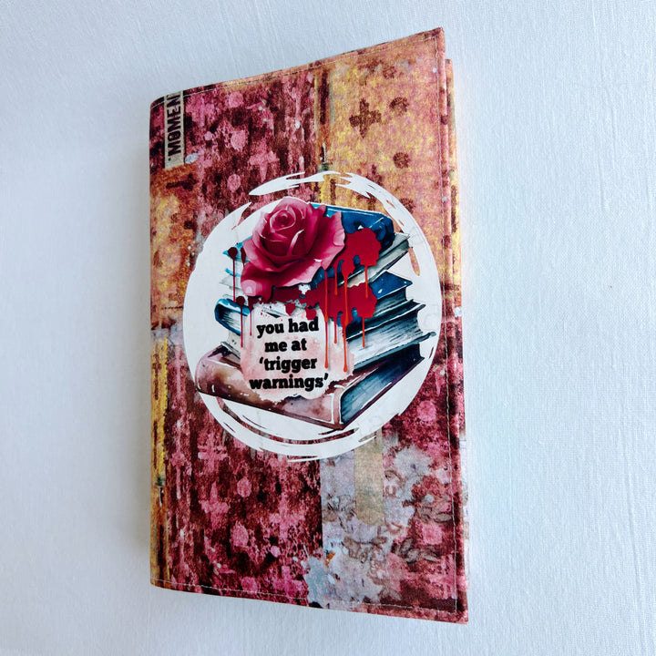 a book covered in a colorful fabric reusable book cover in colors of pinks and orange. A white circle on the front contains an original design with a stack of books, a rose, blood drips and the words, 'you had me at trigger warnings' finishes the look.  Meant for readers of dark romance.