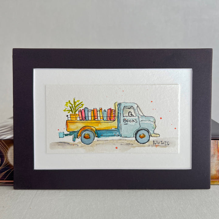 A matted watercolor art print featuring a vintage truck carrying colorful books and a plant, presented in a black frame with a white mat against a neutral background.