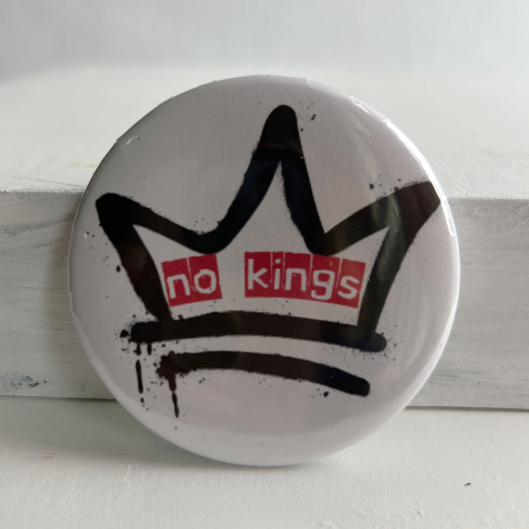 a round, white pinback button with a black crown and  the words, 'no kings' in red.