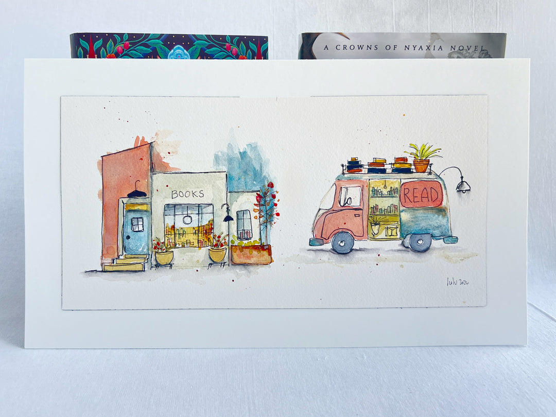 A watercolor art print depicting a cozy bookstore and a retro van with a 'READ' sign, displayed on a white surface with two books behind it.