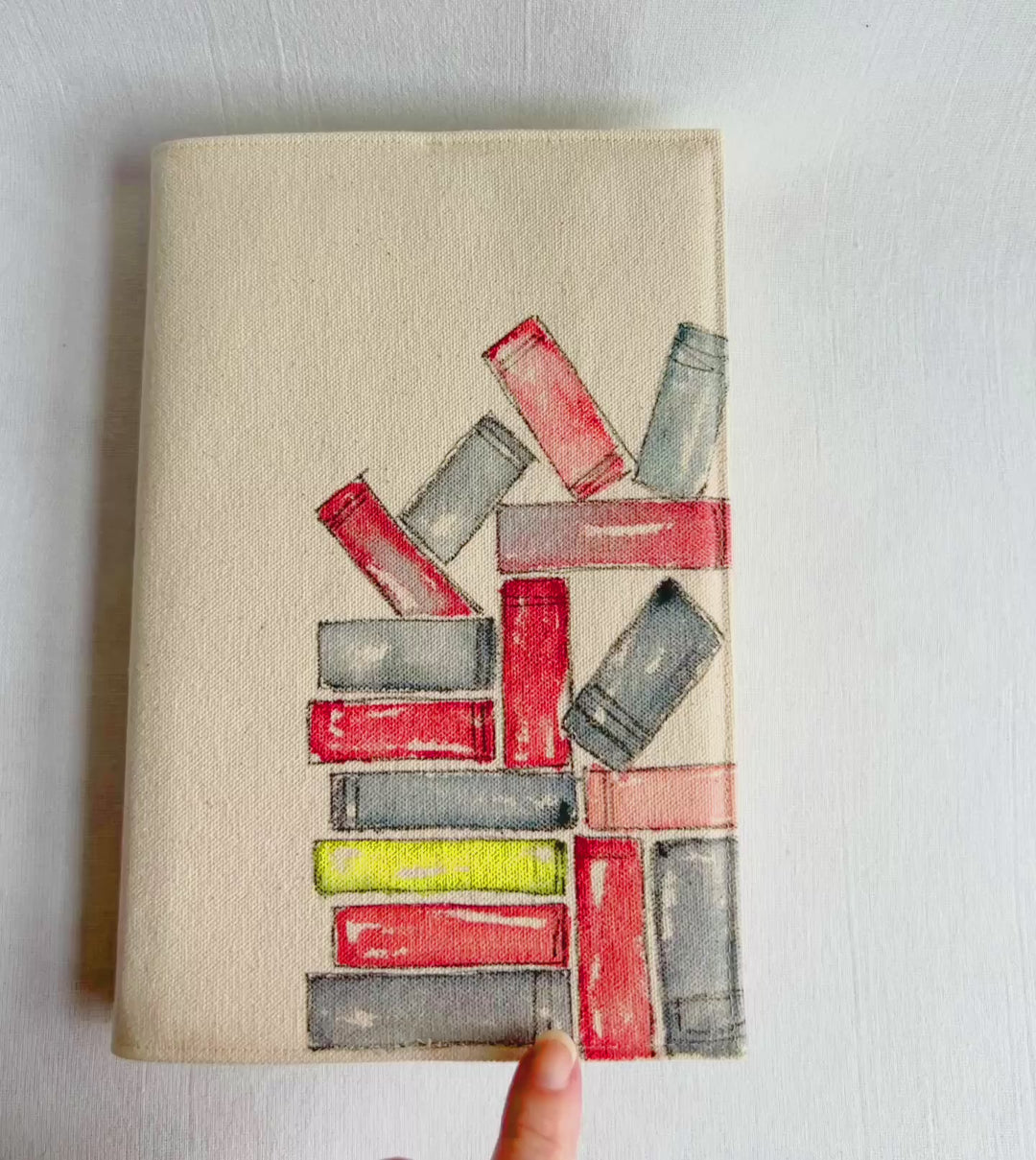 A rectangular canvas book cover featuring a watercolor illustration of stacked books in red, gray, and yellow on a beige background for the outside, and colorful book fabric for the inside opened up to show the inside of the cover