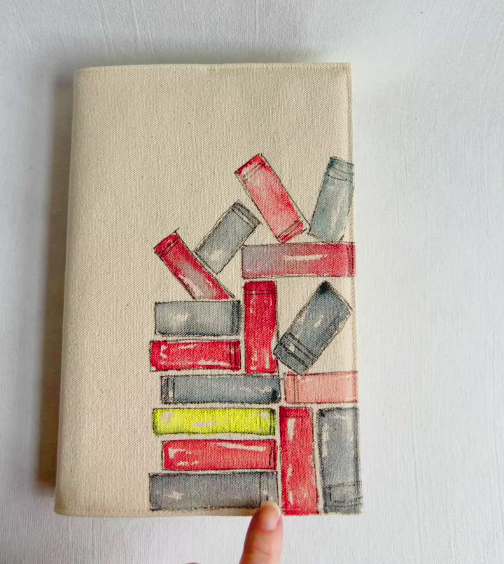A rectangular canvas book cover featuring a watercolor illustration of stacked books in red, gray, and yellow on a beige background for the outside, and colorful book fabric for the inside opened up to show the inside of the cover