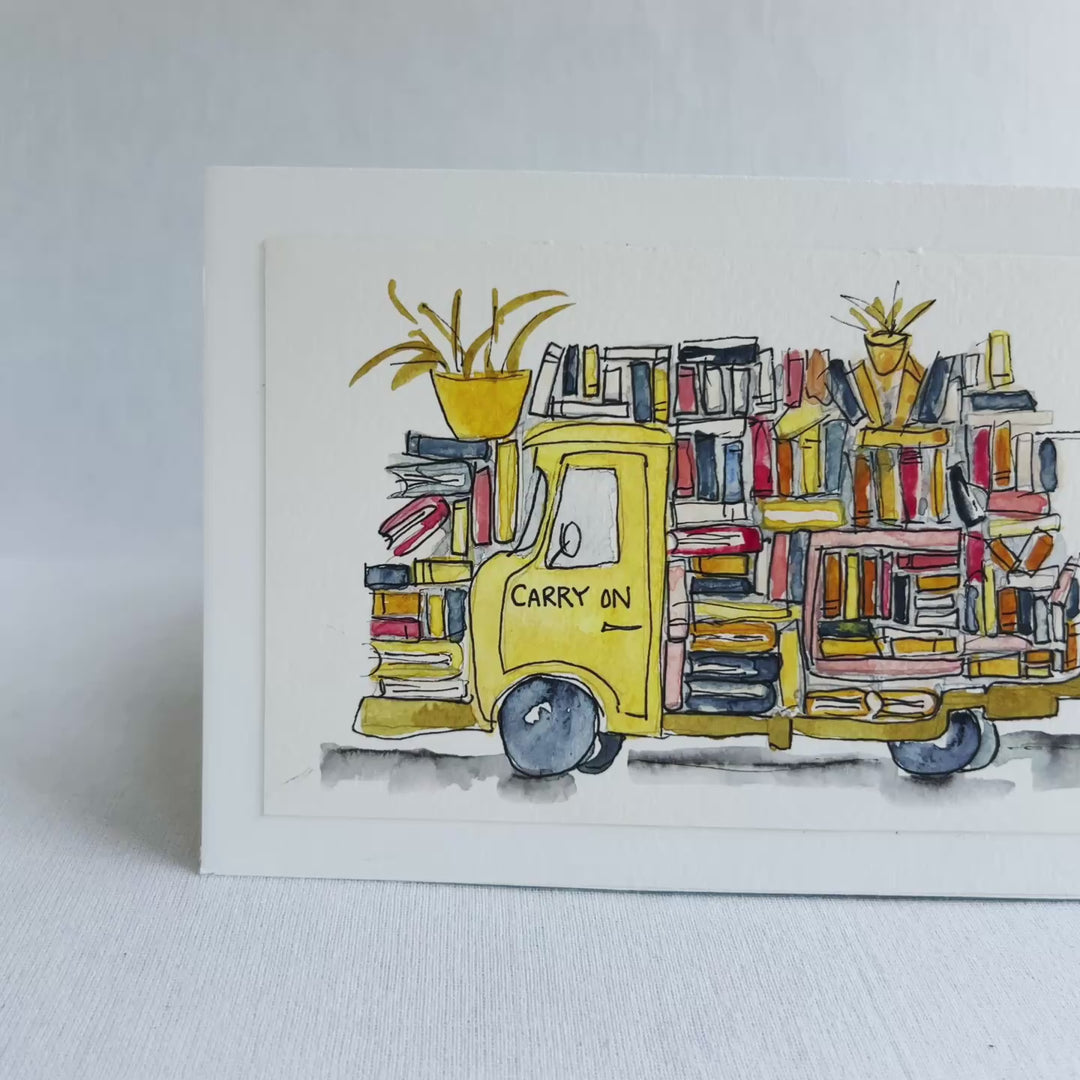 A rectangular watercolor painting of two yellow book trucks stacked and overloaded with books and potted plants, plus a wonky book light, all displayed on top of a decorative pink floral book  against a white background.