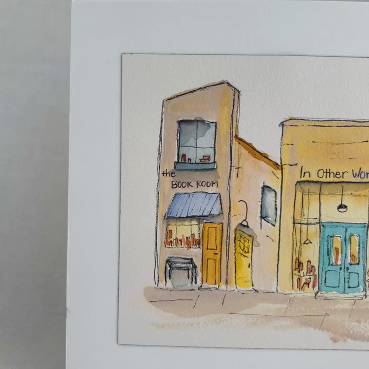 A watercolor art print depicting four stone bookstore buildings, presented with a wide white mat board against a neutral gray background.