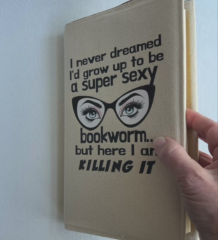 a short video  showing  the inside of a book covered in a reusable linen-color canvas book cover, with the words, 'I never dreamed I'd grow up to be a super sexy bookworm, but here I am killing it.' A pair of eyes with long lashes behind cat eye glasses completes the design.
